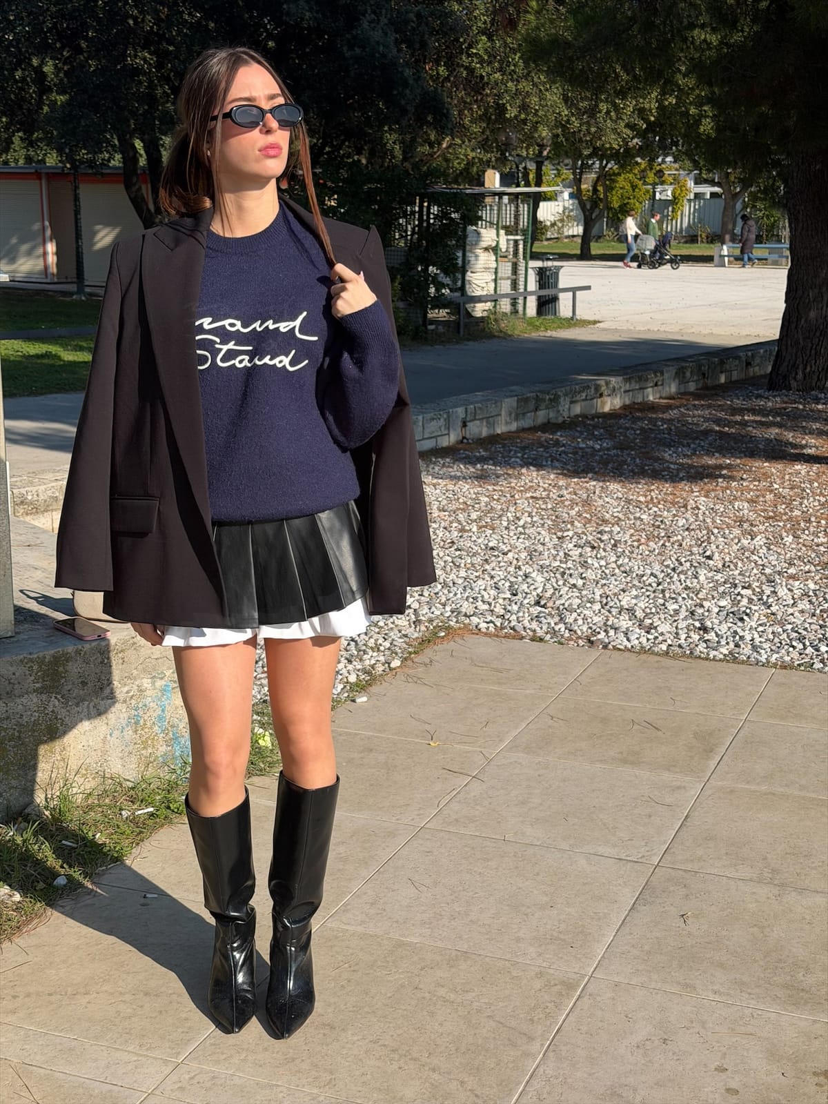 Ena walking outdoors in a navy sweater and tall boots, a blazer over her arm